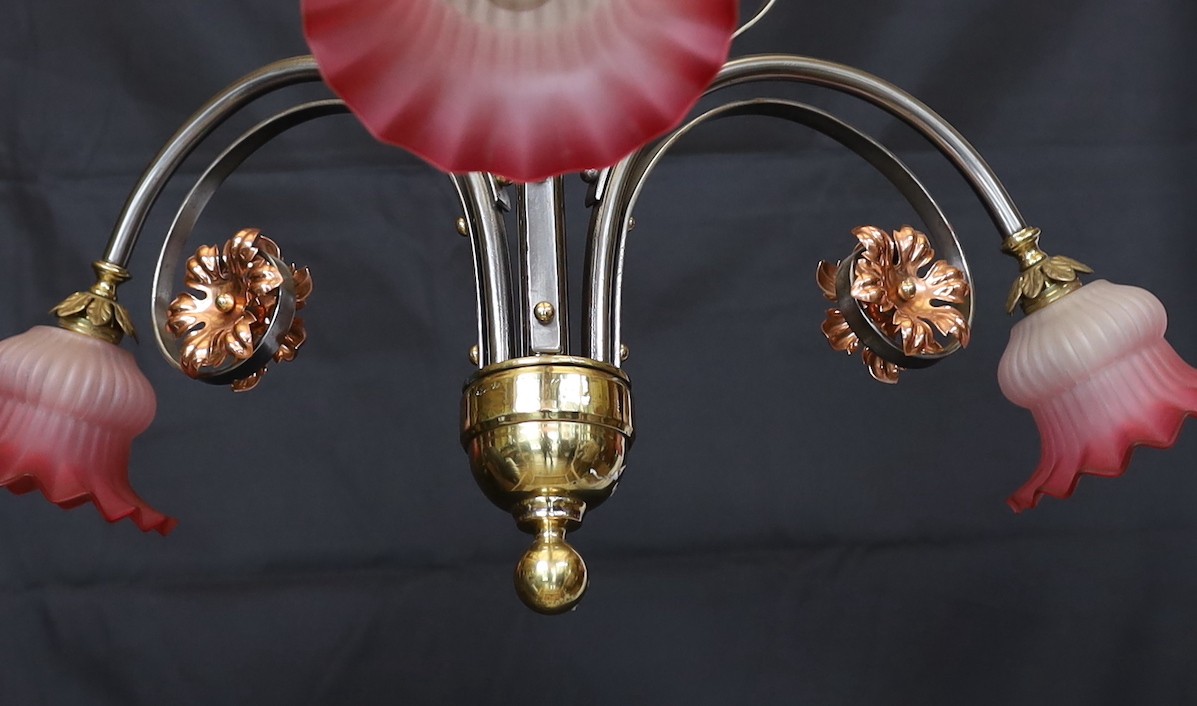 An English Arts & Crafts wrought iron brass and copper three light electrolier with pink tinted opaque glass shades, height 60cm. width 66cm